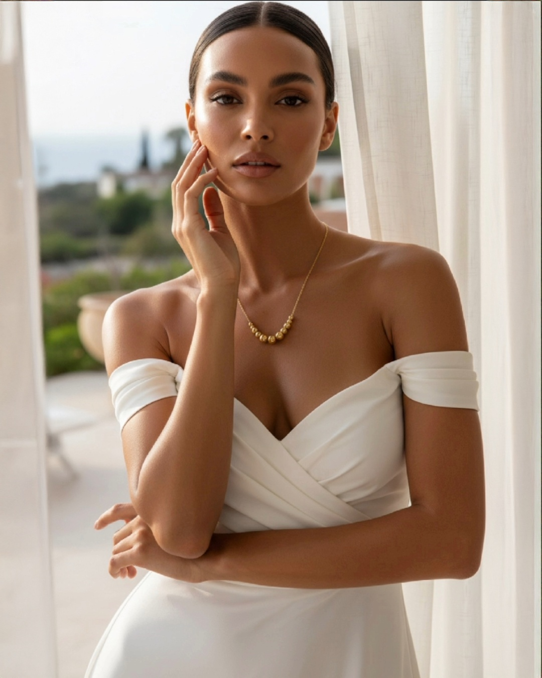 Elegantes Portrait einer stilvollen Frau in einem schulterfreien weißen Kleid vor hellem, mediterranem Hintergrund. Sie trägt eine filigrane Goldhalskette mit mehreren polierten Goldkugeln als zentrales Element. Das warme Licht betont ihre glatte Haut, die feinen Gesichtszüge und die luxuriöse, minimalistische Ästhetik des Schmucks. Die Szene wirkt hochwertig, ruhig und exklusiv.