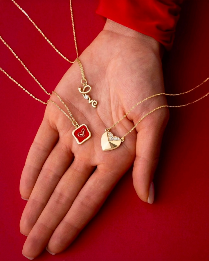 Nahaufnahme einer Hand mit drei filigranen 18k Gelbgold-Halsketten auf rotem Hintergrund, jede mit einem romantischen Anhänger: Schriftzug „Love“ mit kleinem Stein, rotes Emaille-Herz im Quadrat und goldenes Herz mit teils pavégefassten Steinen.