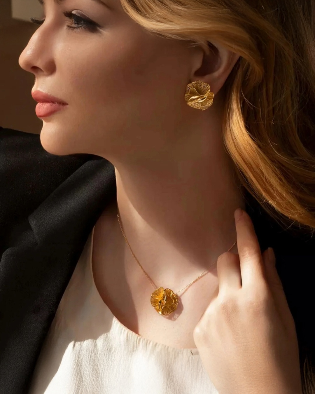 Elegante Frau in warmem Seitenlicht mit goldfarbenen Ohrringen und passender 18k-ähnlicher Halskette. Beide Schmuckstücke haben eine organische, blütenartige Struktur mit glänzender und leicht gefalteter Oberfläche. Die Frau trägt ein helles Top und einen schwarzen Blazer, ihr Kopf ist leicht zur Seite gedreht, wodurch die Details des Schmucks gut sichtbar sind.