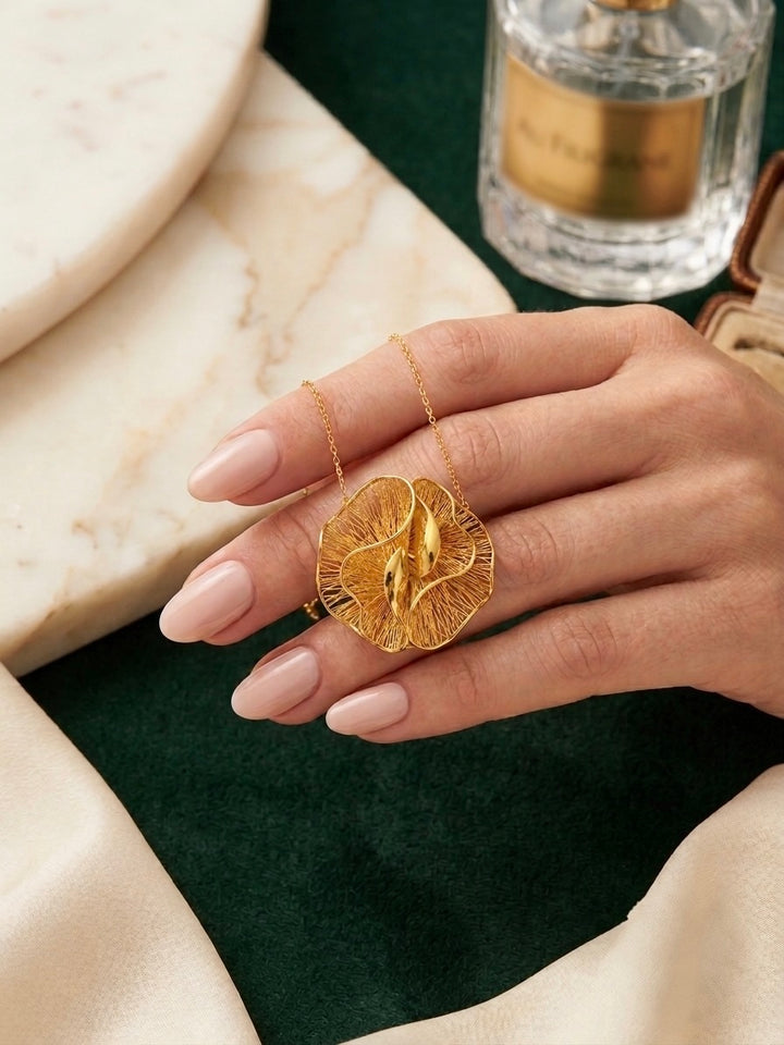 Nahaufnahme einer eleganten Halskette aus 18 Karat Gelbgold, präsentiert auf einer Hand mit gepflegten, dezent lackierten Nägeln. Der filigrane Anhänger besticht durch sein organisch-florales Design mit fein durchbrochenen Strukturen und geschwungenen, hochglanzpolierten Goldelementen, die das Licht wunderschön reflektieren. Die Aufnahme vermittelt die tatsächliche Grösse des Schmuckstücks und unterstreicht seine luxuriöse, moderne Ausstrahlung. Im Hintergrund sorgen Marmor, ein Parfumflakon und weicher Sto