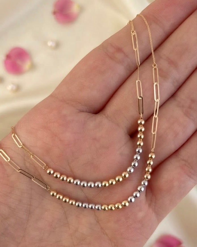 Nahaufnahme einer filigranen zweifarbigen Goldkette auf einer Hand. Die Kette kombiniert längliche, geometrische Kettenglieder mit kleinen gold- und silberfarbenen Kugeln in einem eleganten, modernen Design.