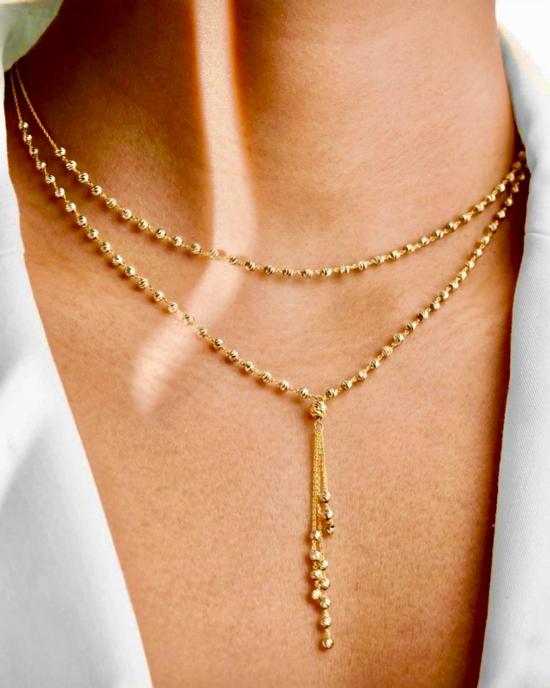 Elegante Y-Halskette aus 18 K Gelbgold mit feinen Goldkugeln, zweireihig gearbeitet und mit filigranem, herabhängendem Kettenelement, getragen am Hals einer Frau vor hellem Hintergrund.