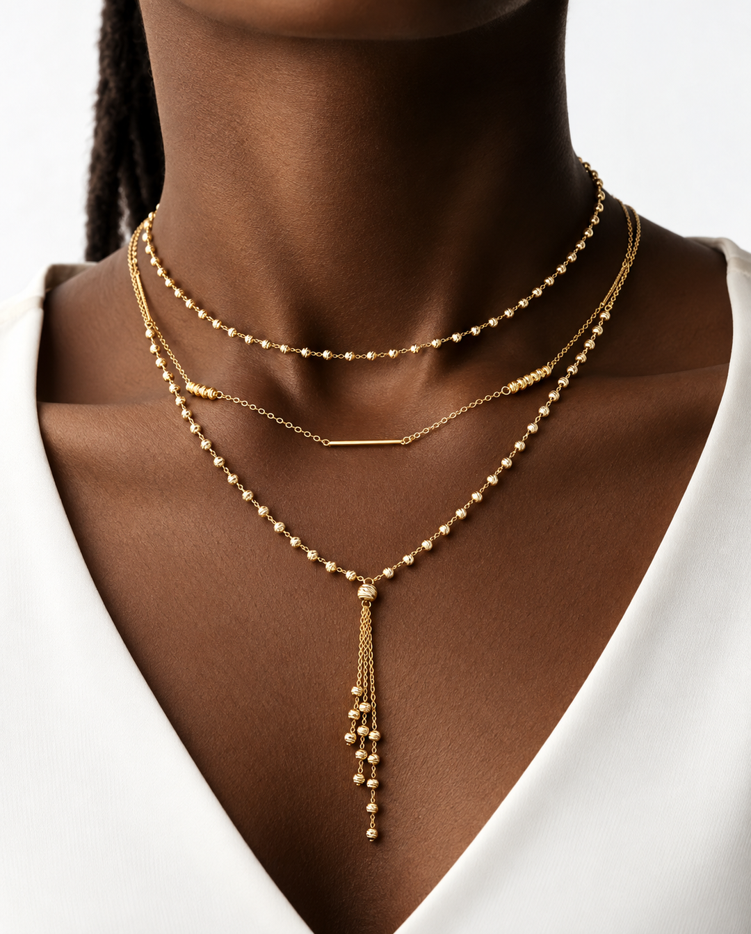 Nahaufnahme eines eleganten, dreilagigen 18k Gelbgold-Halsketten-Ensembles auf dunkler Haut. Die obere Kette besteht aus kleinen, polierten Goldkügelchen, die mittlere aus einer feinen Gliederkette mit schlichtem Goldstab, und die untere Kette aus filigranen Goldperlen mit einem langen, herabfallenden Anhänger aus zarten Goldketten. Das Ensemble wirkt luxuriös, modern und perfekt abgestimmt auf das weiße Oberteil.