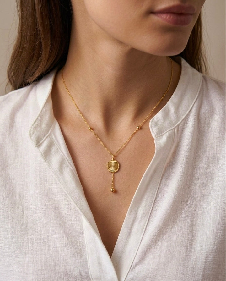 Nahaufnahme einer Frau in weisser Bluse mit einer feinen goldenen Halskette. Die Kette trägt einen runden, strukturierten Anhänger mit einem filigranen Kettenelement und einer kleinen goldenen Kugel als Abschluss.