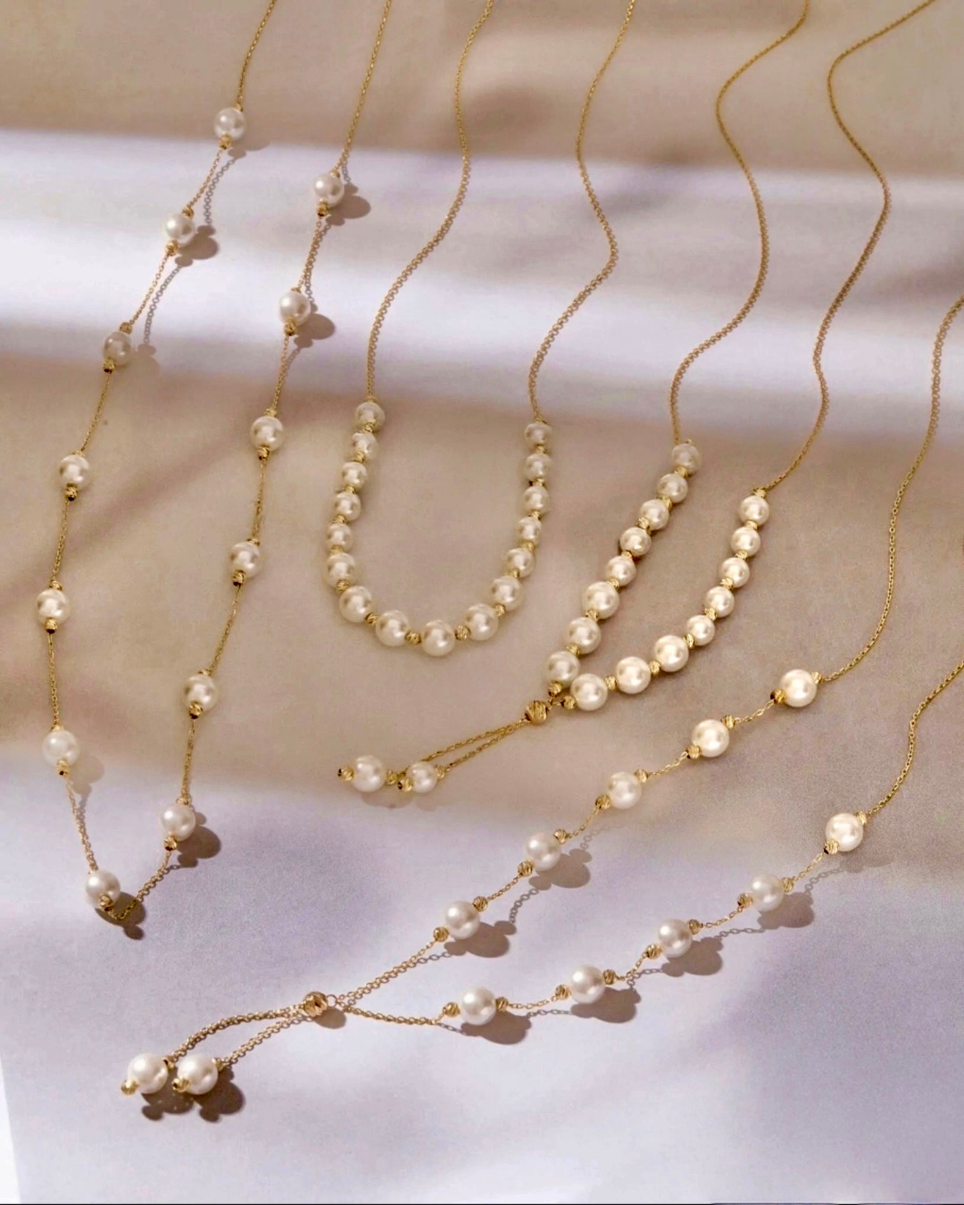 Elegante Halsketten aus 18k Gelbgold mit weißen Perlen, in verschiedenen Längen und Anordnungen, die Perlen wie tanzend auf den feinen Goldketten verteilt, sanft im Licht schimmernd.