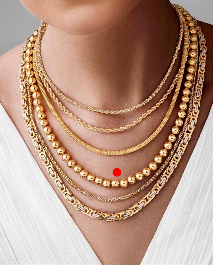 Goldperlenkette aus 18k Gelbgold im Layering-Look getragen, kombiniert mit weiteren Ketten auf heller Haut und weißem Oberteil, markiert für Fokus