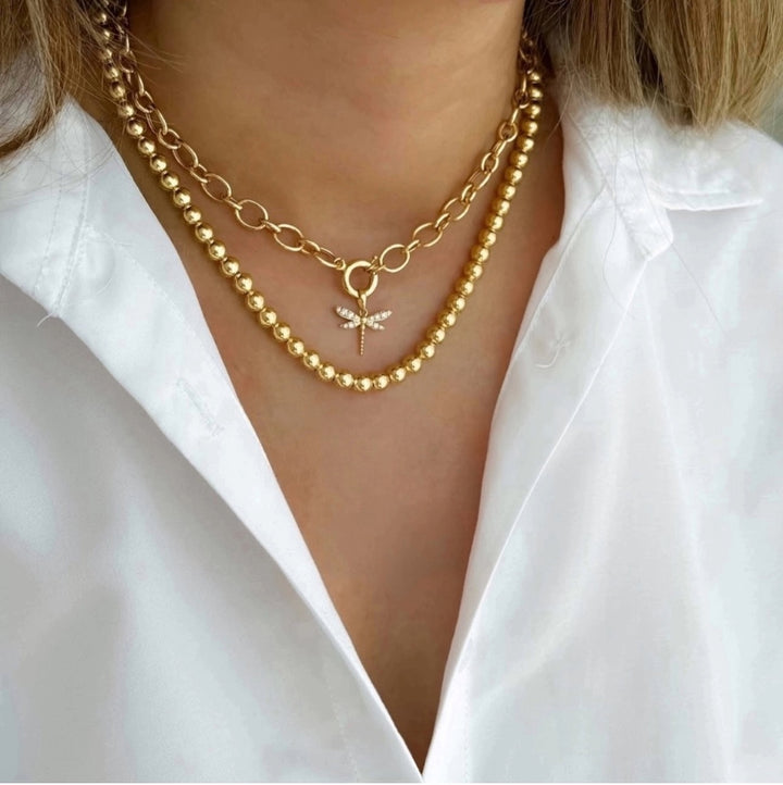 Nahaufnahme eines Halses mit zwei übereinander getragenen Halsketten aus 18k Gelbgold. Die obere Kette besteht aus großen, ovalen Gliedern mit einem dekorativen Ringverschluss, an dem ein Libellenanhänger mit funkelnden Steinen hängt. Die untere Kette ist eine gleichmäßig runde Goldperlenkette mit polierter Oberfläche. Getragen zu einer weißen Bluse mit offenem Kragen.