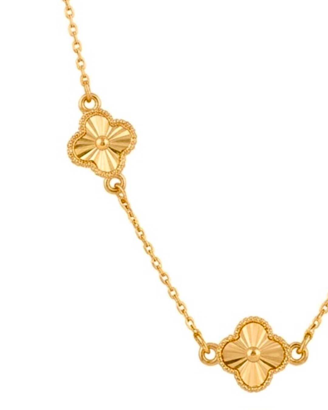 Detailaufnahme einer eleganten Halskette aus 18 Karat Gelbgold mit zwei filigran gestalteten Kleeblatt-Elementen. Die glänzenden Anhänger zeigen ein strahlenförmiges Muster mit feiner Perlenrand-Verzierung und sind durch eine zarte Goldkette verbunden. Der Schmuck ist vor hellem Hintergrund fotografiert und betont die hochwertige Verarbeitung sowie den warmen Goldglanz.