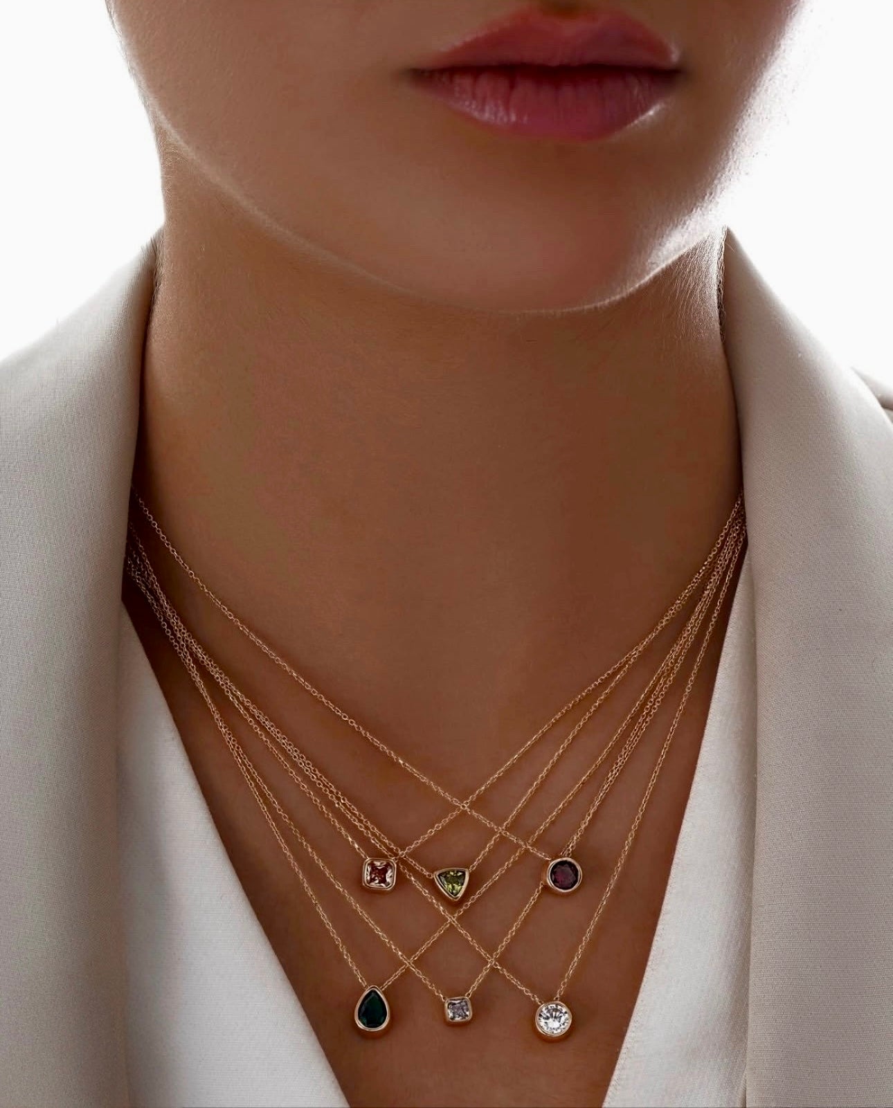 Nahaufnahme eines Halses mit sechs filigranen Halsketten aus 18k Gelbgold, die in Lagen übereinander getragen werden. Jede Kette trägt einen einzelnen Edelsteinanhänger in verschiedenen Formen und Farben, darunter ein grüner, dreieckiger Stein („Peymân-e Sabz“ – Das grüne Versprechen) in der Mitte. Getragen zu einem eleganten, weißen Blazer mit tiefem Ausschnitt.
