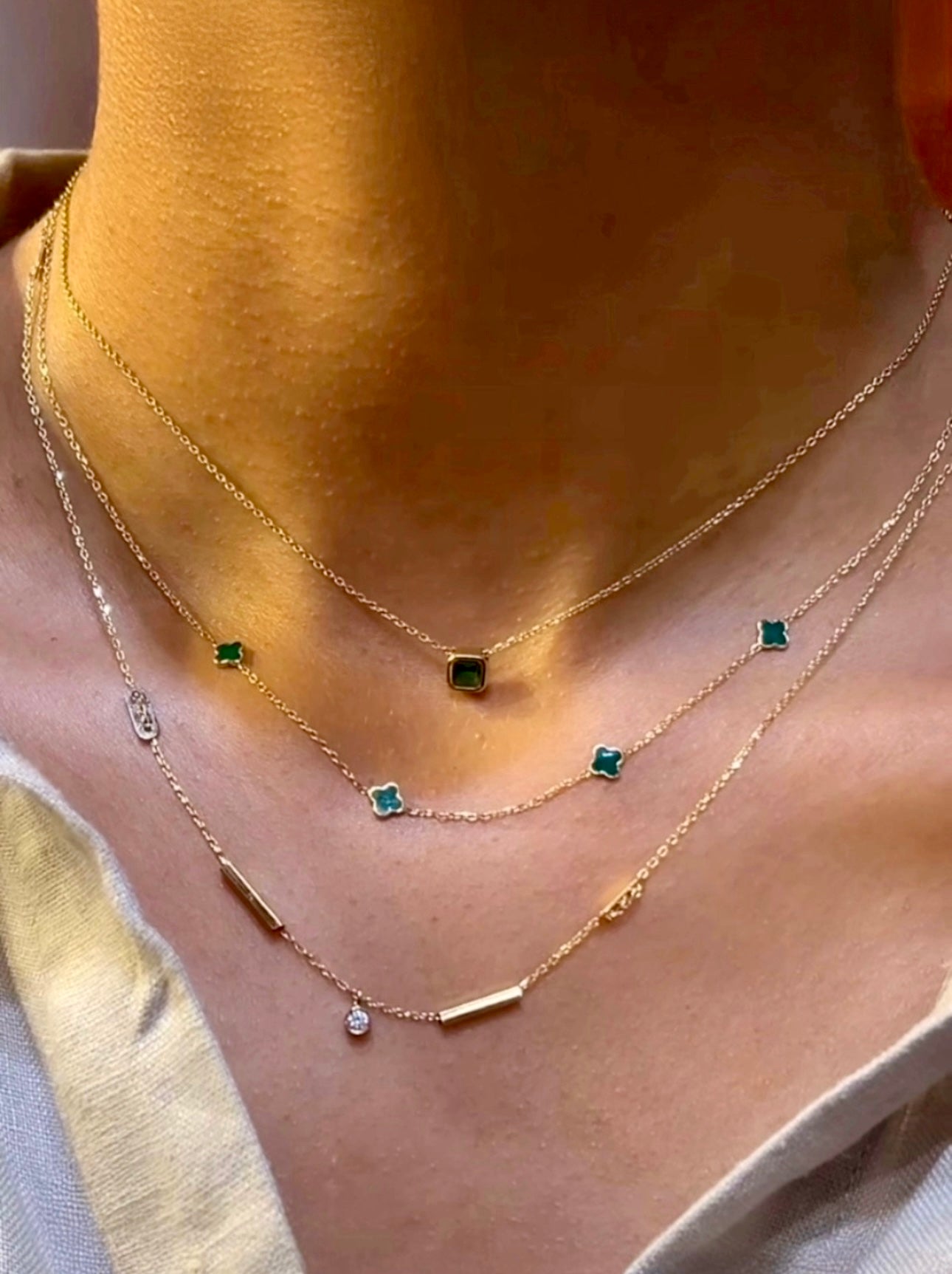 Nahaufnahme eines Halses mit drei filigranen Halsketten aus 18k Gelbgold. Die mittlere Kette („Peymân-e Sabz“ – Das grüne Versprechen) ist mit mehreren kleinen, kleeblattförmigen Anhängern in sattem Smaragdgrün besetzt. Darüber eine kurze Kette mit einem einzelnen quadratischen grünen Edelsteinanhänger, darunter eine längere Kette mit schlichten Goldzylindern und einem einzelnen klaren Steinanhänger. Getragen zu einem hellen, tief ausgeschnittenen Oberteil.