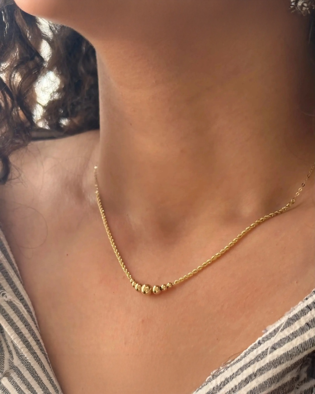 Nahaufnahme eines weiblichen Halses mit einer zarten 18k-Goldkette. Die Kette hat mehrere hochglanzpolierte Goldkugeln in abgestufter Größe im Zentrum und liegt elegant auf dem Schlüsselbein. Warmes Licht betont Glanz und Struktur des Schmucks.