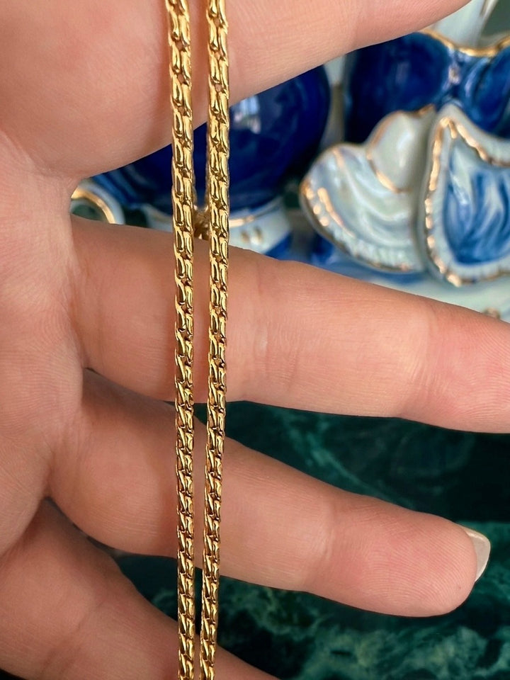 Nahaufnahme einer filigranen Schlangenkette aus 18 Karat Gelbgold mit strukturierter Oberfläche. Die Kette wird zwischen den Fingern gehalten und zeigt ihr elegantes, flexibles Gliederdesign im Detail. Im Hintergrund sind unscharfe blau-weisse Keramikdekorationen zu sehen, welche den warmen Goldton besonders hervorheben. Die hochwertige Verarbeitung und der edle Glanz verleihen der Halskette eine zeitlose, luxuriöse Ausstrahlung.