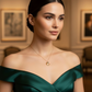 Elegante Frau mit zurückgestecktem dunklem Haar trägt ein schulterfreies, smaragdgrünes Kleid und eine feine Halskette mit einem kleinen goldenen Anhänger, aufgenommen in einem stilvollen, warm beleuchteten Galerieraum.