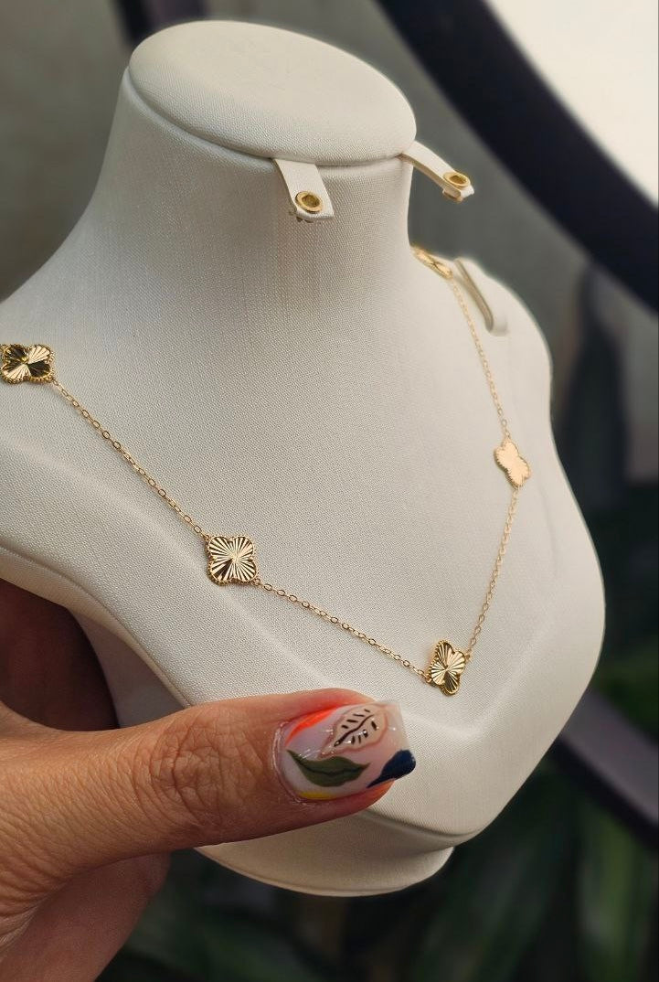 Eine feine 18k-Gelbgold-Halskette mit kleinen, quadratischen und kleeblattförmigen Zierelementen auf einem weißen Schmuckbüstenständer. Im Vordergrund ist eine Hand mit bunter Nail-Art leicht sichtbar, die den unteren Teil der Büste hält.