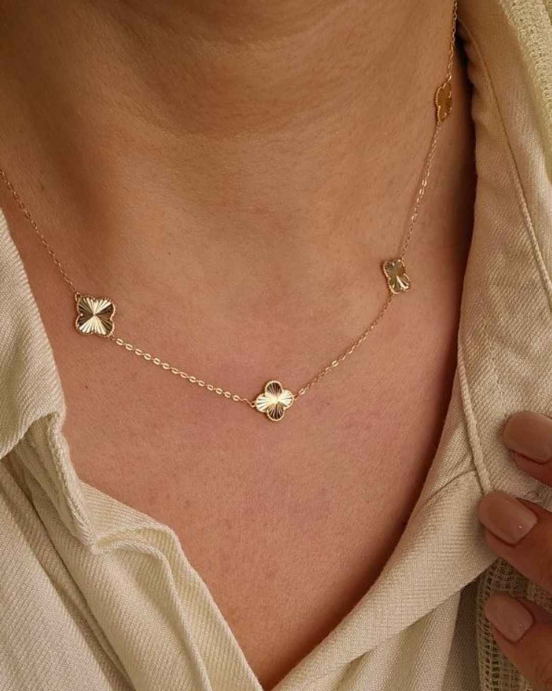 Feine 18k-Gelbgold-Halskette mit drei strukturierten vierblättrigen Ornament-Elementen, getragen nah am Hals auf beiger Bluse; warmes, natürliches Licht betont Glanz und Details.