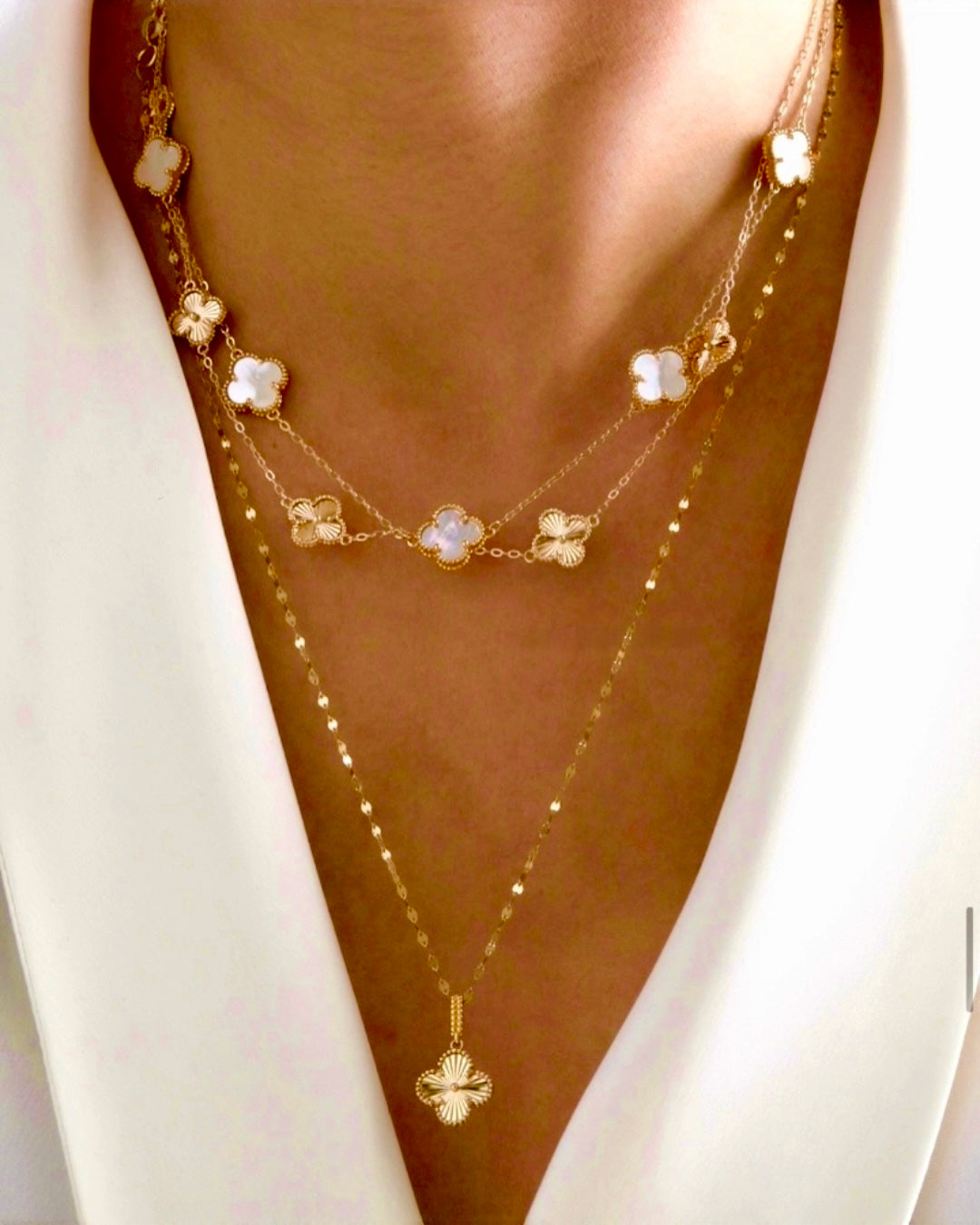 Mehrreihige Halskette aus Gelbgold 18k mit feinen Kettenlagen und dekorativen Kleeblatt-Elementen, teils mit weißer Emaille, elegant am Dekolleté getragen; lange Kette mit filigranem Kleeblatt-Anhänger, stilvoll kombiniert zu einem luxuriösen Layering-Look.