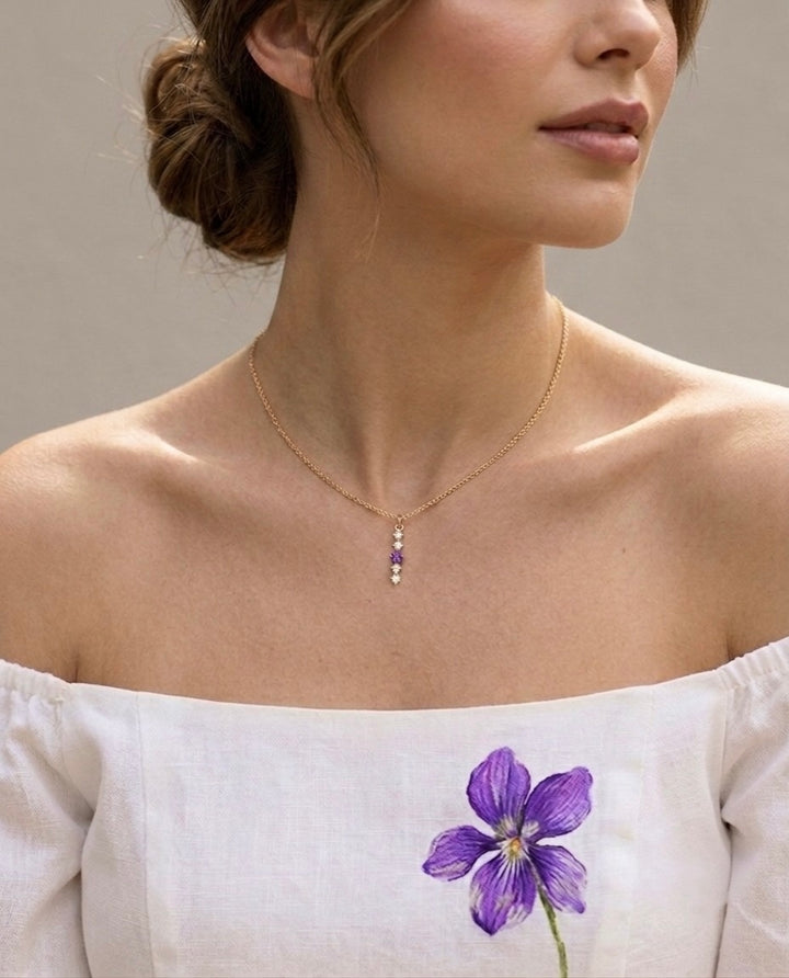 Nahaufnahme einer Frau mit locker hochgestecktem Haar und schulterfreiem weissem Oberteil mit einer violetten Blüte. Um ihren Hals trägt sie eine filigrane Gelbgold-Halskette mit einem eleganten vertikalen Anhänger aus fünf funkelnden Steinen. In der Mitte leuchtet ein violetter Edelstein, eingerahmt von vier klaren Steinen, wodurch ein harmonischer und femininer Farbakzent entsteht. Der neutrale Hintergrund und das weiche Licht lenken den Fokus auf den zarten Schmuck und unterstreichen dessen edle Ausstrah
