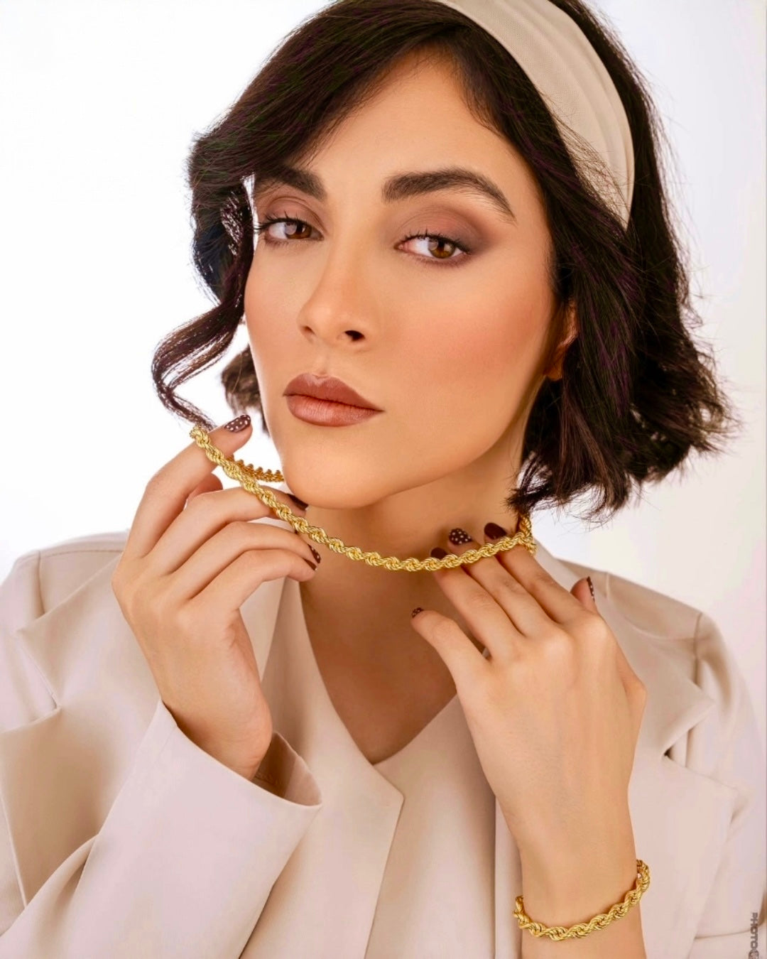 Elegante Frau trägt eine geflochtene Halskette und ein passendes Armband aus poliertem Gelbgold, stilvoll inszeniert vor hellem Hintergrund mit natürlichem Make-up und cremefarbenem Outfit.