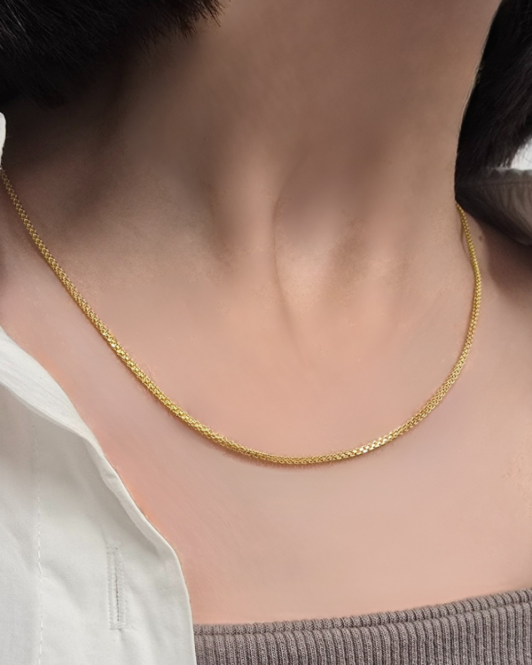 Nahaufnahme eines Halses mit einer feinen, gleichmäßig gearbeiteten 18k-Goldkette. Die Kette liegt elegant am Schlüsselbein an, wirkt glatt, glänzend und dezent. Der Hintergrund zeigt neutrale Kleidung, wodurch das Gold stärker hervortritt.