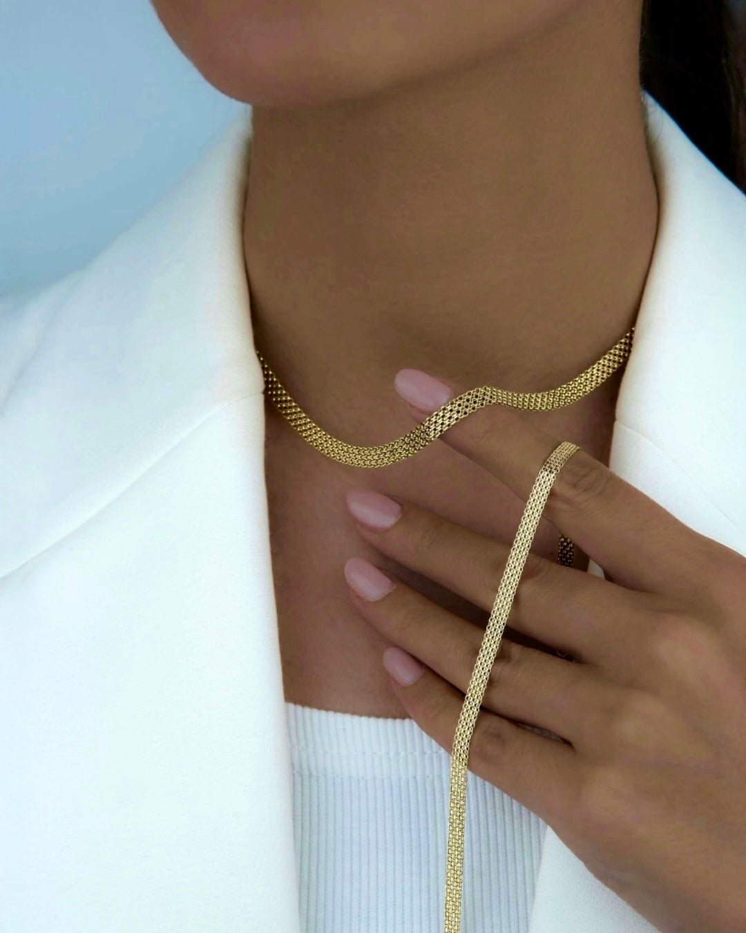 Detailaufnahme einer Frau mit einer flach gearbeiteten Herringbone-Halskette aus 18k Gelbgold, die sanft in einer geschwungenen Linie am Hals anliegt; edler Goldglanz, kombiniert mit einem hellen Blazer und natürlichem Styling.