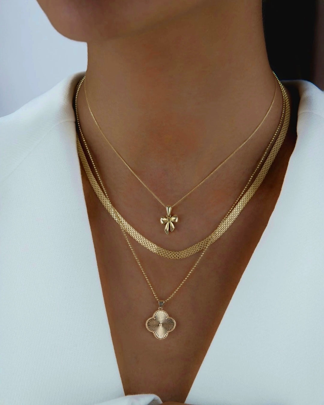 Stilvolle Layering-Aufnahme mit drei feinen Halsketten aus 18k Gelbgold: eine flach gearbeitete Herringbone-Kette sowie zwei zarte Goldketten mit elegantem Schleifenanhänger und ornamentförmigem Anhänger, getragen zu einem hellen Blazer mit tiefem Ausschnitt.