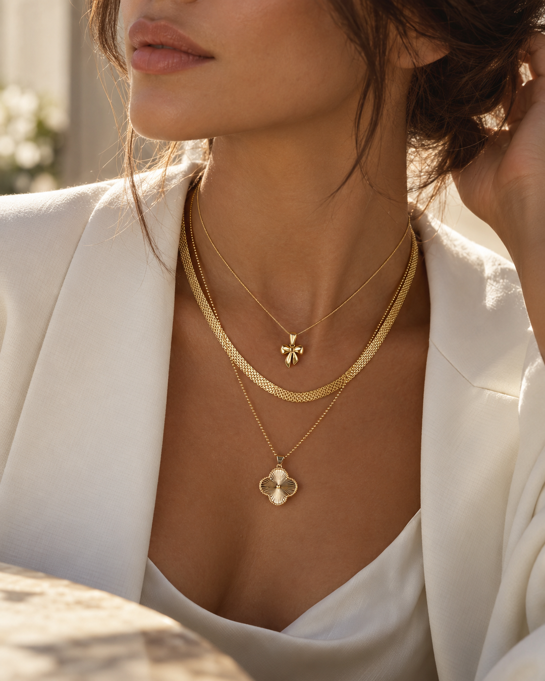 Elegantes Fashion-Editorial mit einer Frau im weissen Blazer und mehreren übereinander getragenen Goldketten mit Schleifen- und floralem Anhänger im warmen Sonnenlicht.