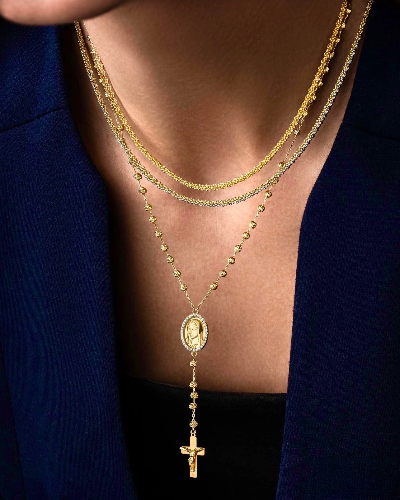 Mehrlagige 18k Goldkette, bestehend aus feinen Gliederketten und einer längeren Rosenkranzkette mit ovalem Medaillon und Kreuzanhänger, getragen über einem dunklen Top und Blazer.