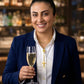 Elegante Frau im dunkelblauen Blazer mit goldenen Knöpfen und weisser Bluse, hält ein Glas Champagner. Sie trägt feinen goldenen Schmuck, darunter Halsketten und Ohrringe, vor einem stilvollen, warm beleuchteten Bar-Hintergrund.