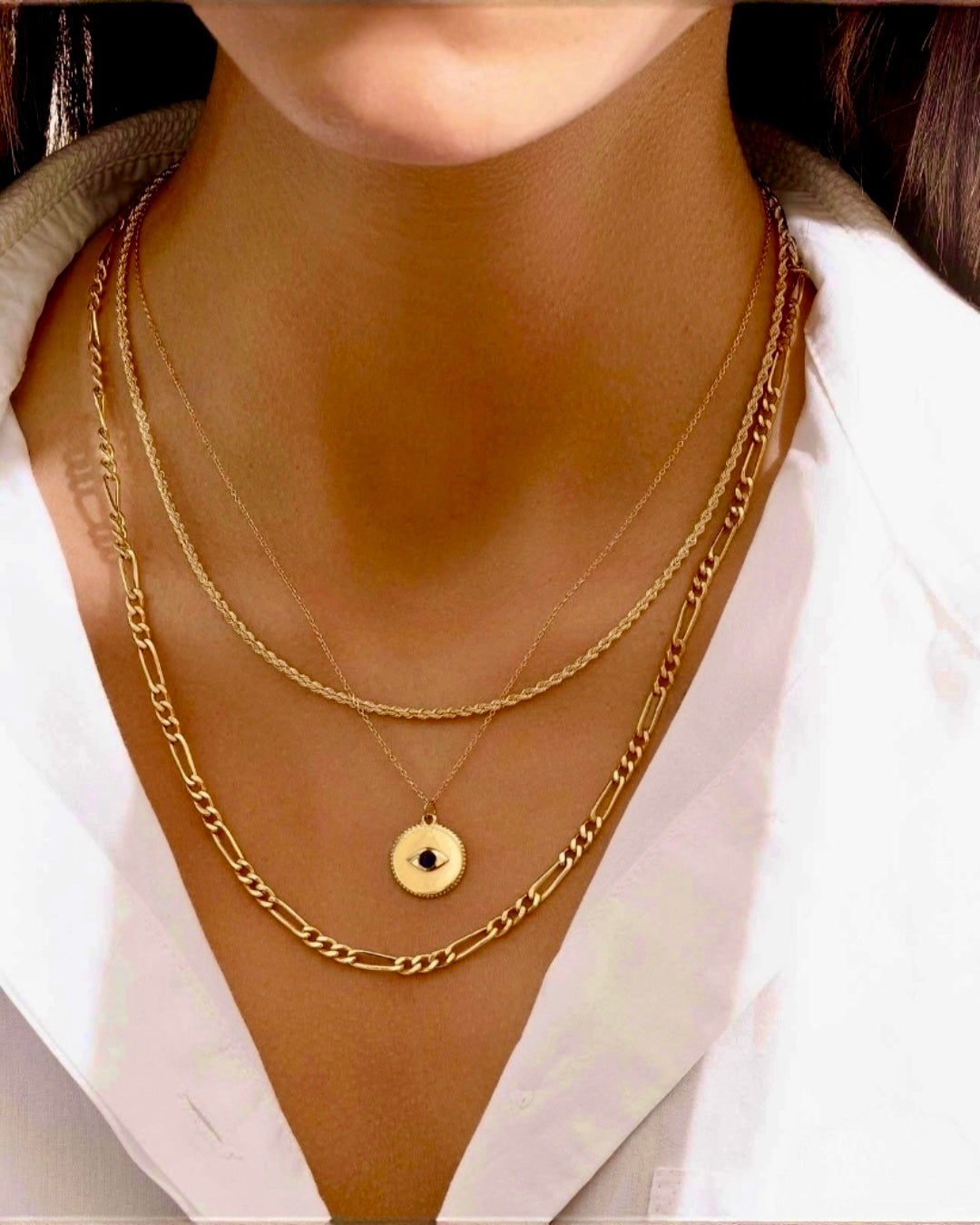 Mehrlagige Goldketten an einem Hals: eine feine Kette mit rundem Anhänger und blauem Stein in Augenform, kombiniert mit einer gedrehten Goldkette und einer markanten Gliederkette, getragen zu einer weißen Bluse.