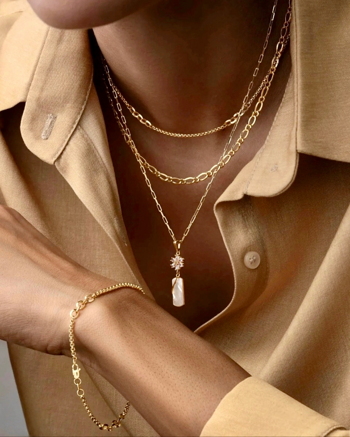 Elegante Frau trägt mehrere Halsketten aus 18k Gelbgold in unterschiedlichen Längen, kombiniert mit einem feinen Armband aus Gold. Die längste Kette zeigt einen Anhänger mit Perlmutt und funkelndem Sternmotiv – luxuriös, modern und raffiniert gestylt über einer sandfarbenen Bluse.