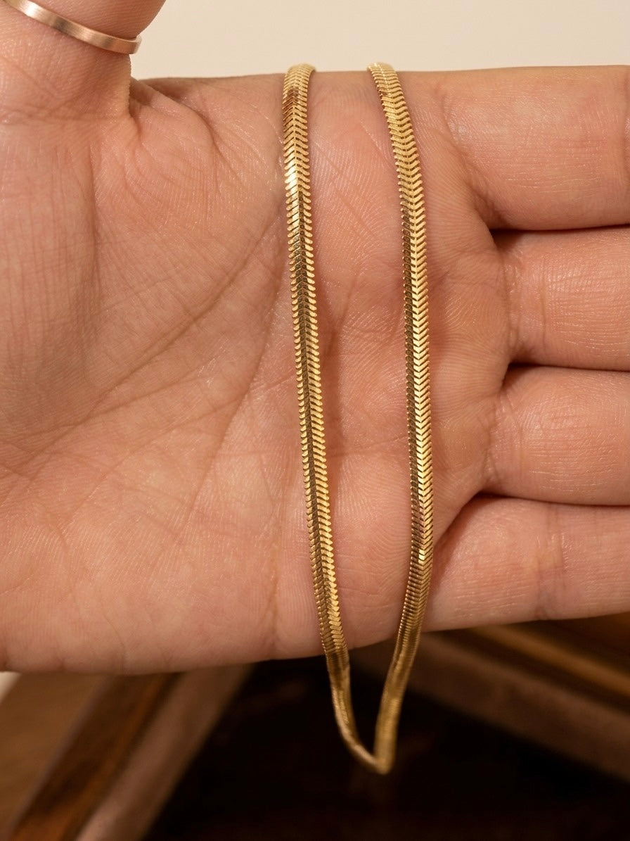Nahaufnahme einer filigranen Fischgrät-Halskette aus 18-karätigem Gelbgold, die auf einer geöffneten Hand präsentiert wird. Die flach anliegenden, fein ineinandergreifenden Glieder bilden das charakteristische V-Muster der Fischgrätkette und reflektieren das Licht mit einem eleganten, seidigen Glanz. Der warme Goldton und die präzise Verarbeitung verleihen der Kette eine zeitlose, edle Ausstrahlung. Im Hintergrund ist eine Holzoberfläche leicht unscharf zu erkennen.