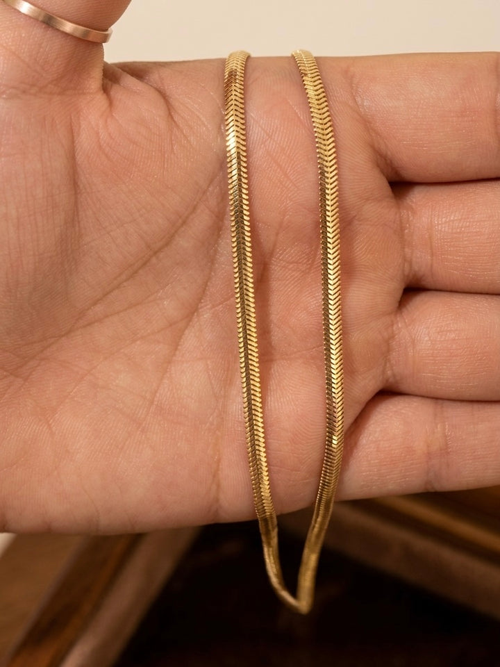 Nahaufnahme einer filigranen Fischgrät-Halskette aus 18-karätigem Gelbgold, die auf einer geöffneten Hand präsentiert wird. Die flach anliegenden, fein ineinandergreifenden Glieder bilden das charakteristische V-Muster der Fischgrätkette und reflektieren das Licht mit einem eleganten, seidigen Glanz. Der warme Goldton und die präzise Verarbeitung verleihen der Kette eine zeitlose, edle Ausstrahlung. Im Hintergrund ist eine Holzoberfläche leicht unscharf zu erkennen.