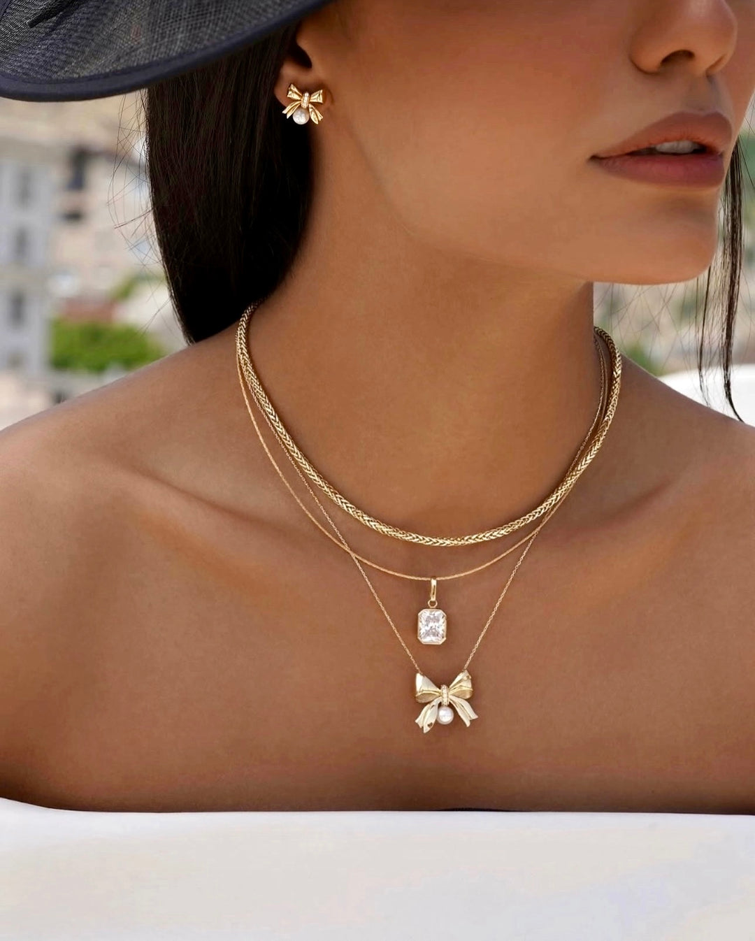 Frau mit eleganter 18k Gelbgold-Halskette im Miro-Design, kombiniert mit zwei weiteren feinen Goldketten, einer rechteckigen Anhängerfassung und einem goldenen Schleifenanhänger mit Perle, passend zu goldenen Schleifenohrringen.