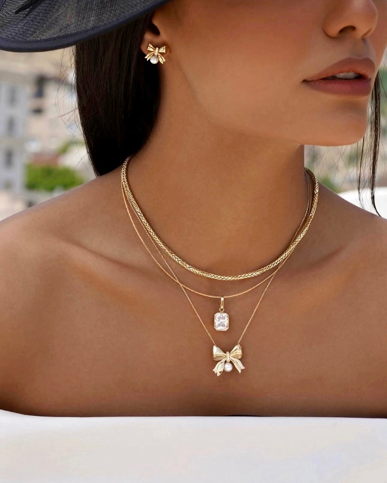 Frau mit eleganter 18k Gelbgold-Halskette im Miro-Design, kombiniert mit zwei weiteren feinen Goldketten, einer rechteckigen Anhängerfassung und einem goldenen Schleifenanhänger mit Perle, passend zu goldenen Schleifenohrringen.