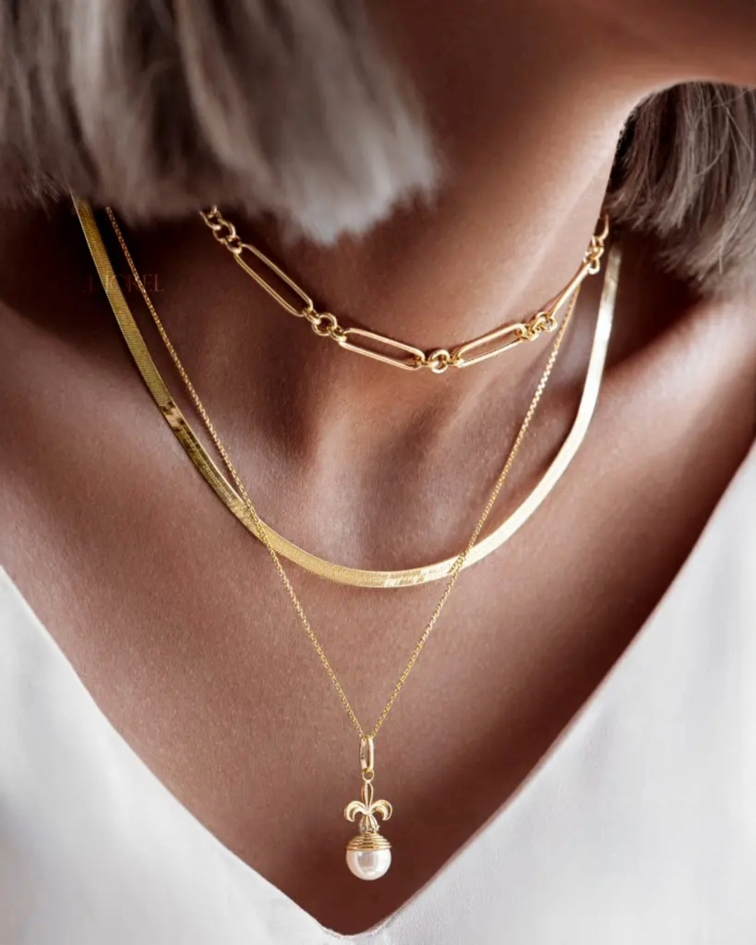 Modell trägt drei übereinander kombinierte 18k-Gelbgold-Halsketten: oben eine Paperclip-Kette, mittig eine flache Fischgrätenkette, unten eine feine Kette mit Perlenanhänger.