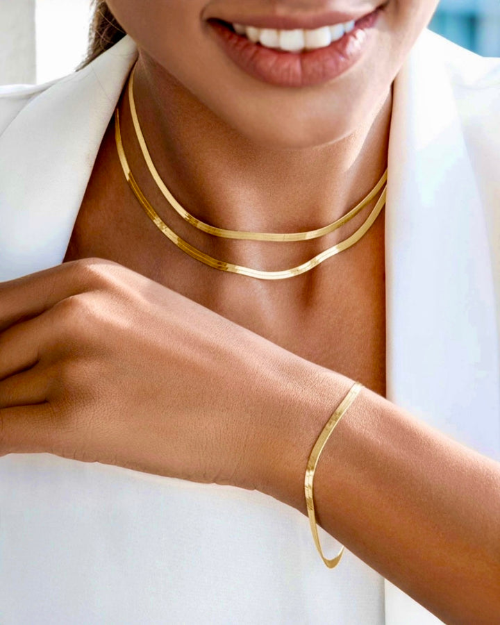 Elegantes Damenarmband und zweireihige Halskette aus poliertem Gelbgold, getragen am Handgelenk und Hals, minimalistisch und zeitlos kombiniert mit weißem Blazer.