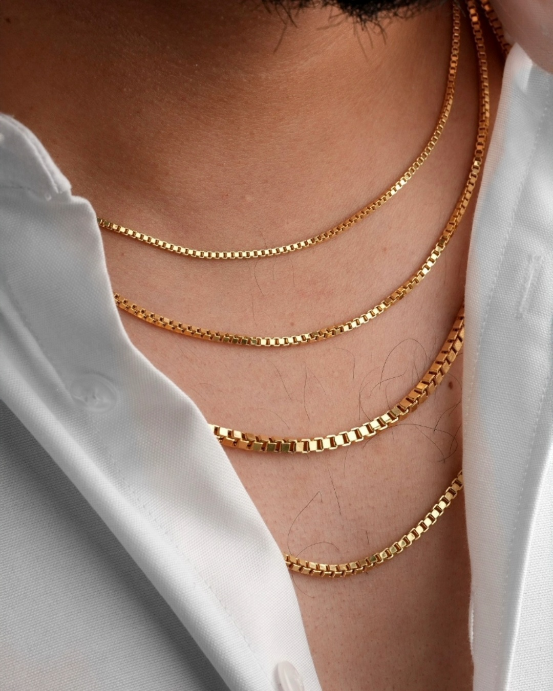Mehrlagig getragene 18k Gelbgold-Halsketten mit eckiger Gliederstruktur (Box Chain) an einem männlichen Hals, kombiniert mit offenem weissen Hemd – minimalistischer, eleganter Goldschmuck mit modernem Layering-Look.