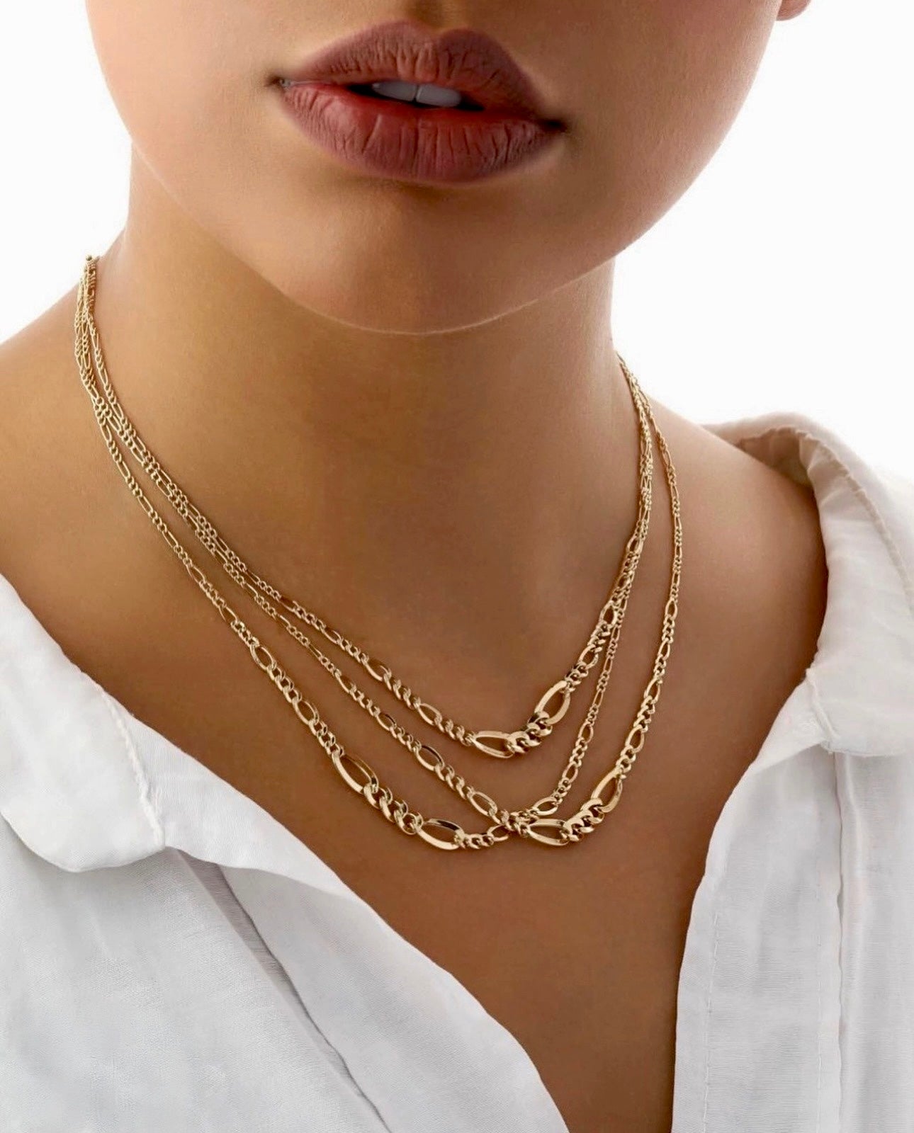 Nahaufnahme einer dreifach gelayerten Halskette aus 18k Gelbgold im Figaro-Conic-Design, getragen über einer weißen, leicht geöffneten Bluse.