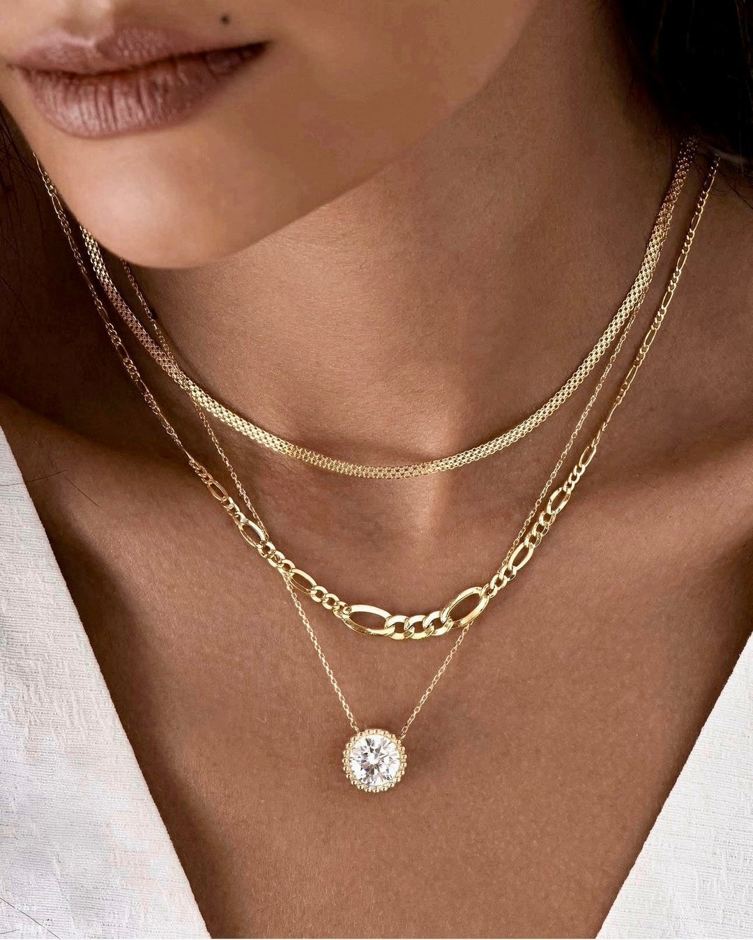 Nahaufnahme eines Halses mit drei übereinander getragenen Halsketten aus 18k Gelbgold. Die mittlere Kette ist eine konische Figaro-Kette mit wechselnden langen und kurzen Gliedern. Darüber liegt eine feine, engmaschige Kette, darunter eine filigrane Kette mit einem großen, ovalen Anhänger, besetzt mit einem funkelnden, klaren Stein. Getragen zu einem tief ausgeschnittenen, weißen Oberteil.