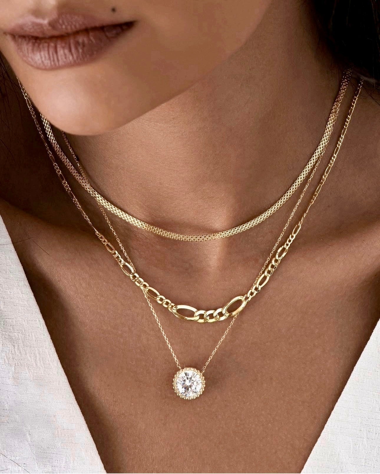 Nahaufnahme eines Halses mit drei übereinander getragenen Halsketten aus 18k Gelbgold. Die mittlere Kette ist eine konische Figaro-Kette mit wechselnden langen und kurzen Gliedern. Darüber liegt eine feine, engmaschige Kette, darunter eine filigrane Kette mit einem großen, ovalen Anhänger, besetzt mit einem funkelnden, klaren Stein. Getragen zu einem tief ausgeschnittenen, weißen Oberteil.