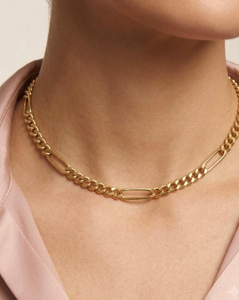 Nahaufnahme des Halses und Dekolletés einer Person mit hellrosafarbener Bluse. Getragen wird eine elegante 18-karätige Gelbgold-Halskette im Figaro-Stil mit einem harmonischen Wechsel aus länglichen ovalen Gliedern und eng aneinanderliegenden Panzergliedern. Die Kette liegt nah am Hals an und wirkt zugleich modern und zeitlos. Der schlichte Hintergrund sowie die weichen Farben der Kleidung lenken den Fokus auf den warmen Glanz des Goldes und das markante Kettendesign.