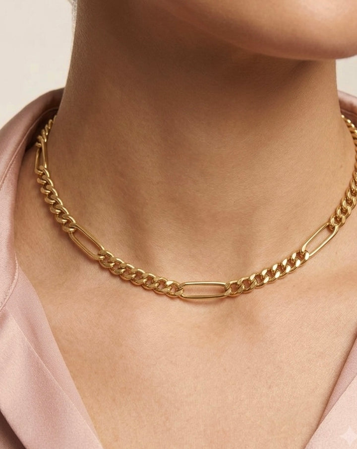 Nahaufnahme des Halses und Dekolletés einer Person mit hellrosafarbener Bluse. Getragen wird eine elegante 18-karätige Gelbgold-Halskette im Figaro-Stil mit einem harmonischen Wechsel aus länglichen ovalen Gliedern und eng aneinanderliegenden Panzergliedern. Die Kette liegt nah am Hals an und wirkt zugleich modern und zeitlos. Der schlichte Hintergrund sowie die weichen Farben der Kleidung lenken den Fokus auf den warmen Glanz des Goldes und das markante Kettendesign.