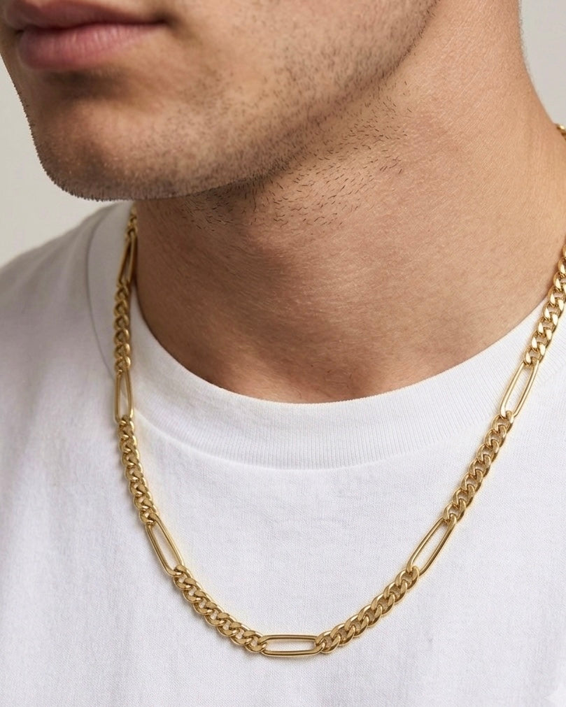 Nahaufnahme des Halses und Unterkiefers eines Mannes mit kurzem Bartschatten, der ein weisses T-Shirt trägt. Um seinen Hals liegt eine massive 18-karätige Gelbgold-Halskette im Figaro-Stil mit einem Wechsel aus länglichen ovalen Gliedern und eng aneinanderliegenden Panzergliedern. Die Kette sitzt locker am Hals und fällt mittig symmetrisch. Der schlichte Hintergrund und das weisse Shirt setzen den warmen Goldton der Kette elegant in Szene.