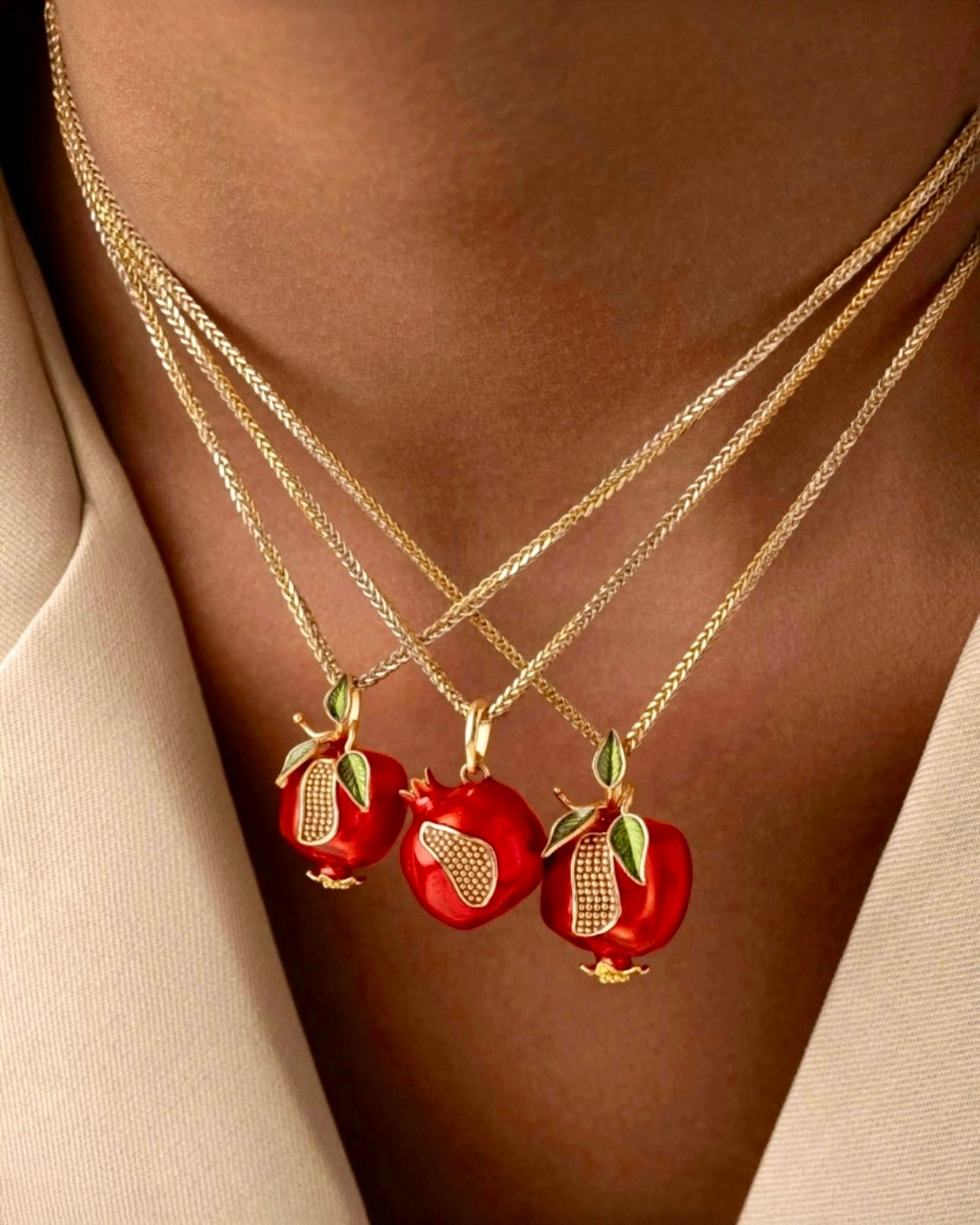 Drei feine Goldketten mit filigranen Granatapfel-Anhängern aus rotem Email und goldenen Details, elegant geschichtet am Dekolleté vor hellem Outfit.