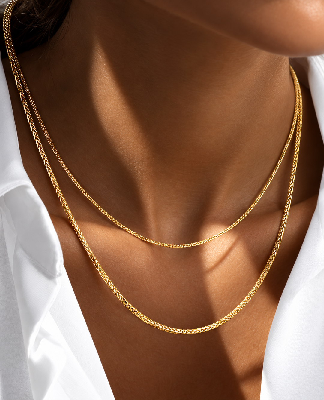 Nahaufnahme eines Halses mit zwei filigranen, glänzenden Miroketten aus 18k Gelbgold und Weissgold in feiner Flechtstruktur, getragen über einem weißen, leicht geöffneten Hemd.