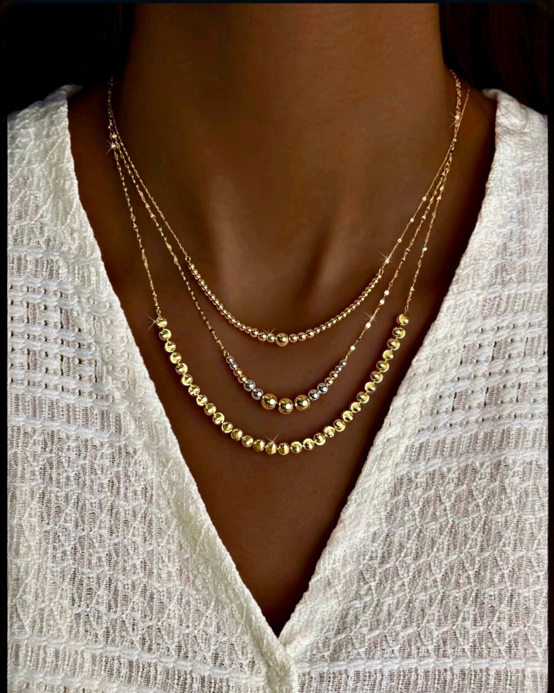 Mehrlagige 18k-Goldhalsketten mit polierten Goldkugeln in abgestuften Längen, elegant am Hals getragen, auf heller Textiloberfläche, Nahaufnahme.