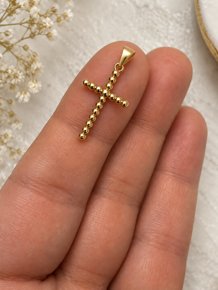 Detailaufnahme des Kreuzanhängers aus 18K Gelbgold mit polierten Goldkugeln und Anhängeröse auf einem Finger.