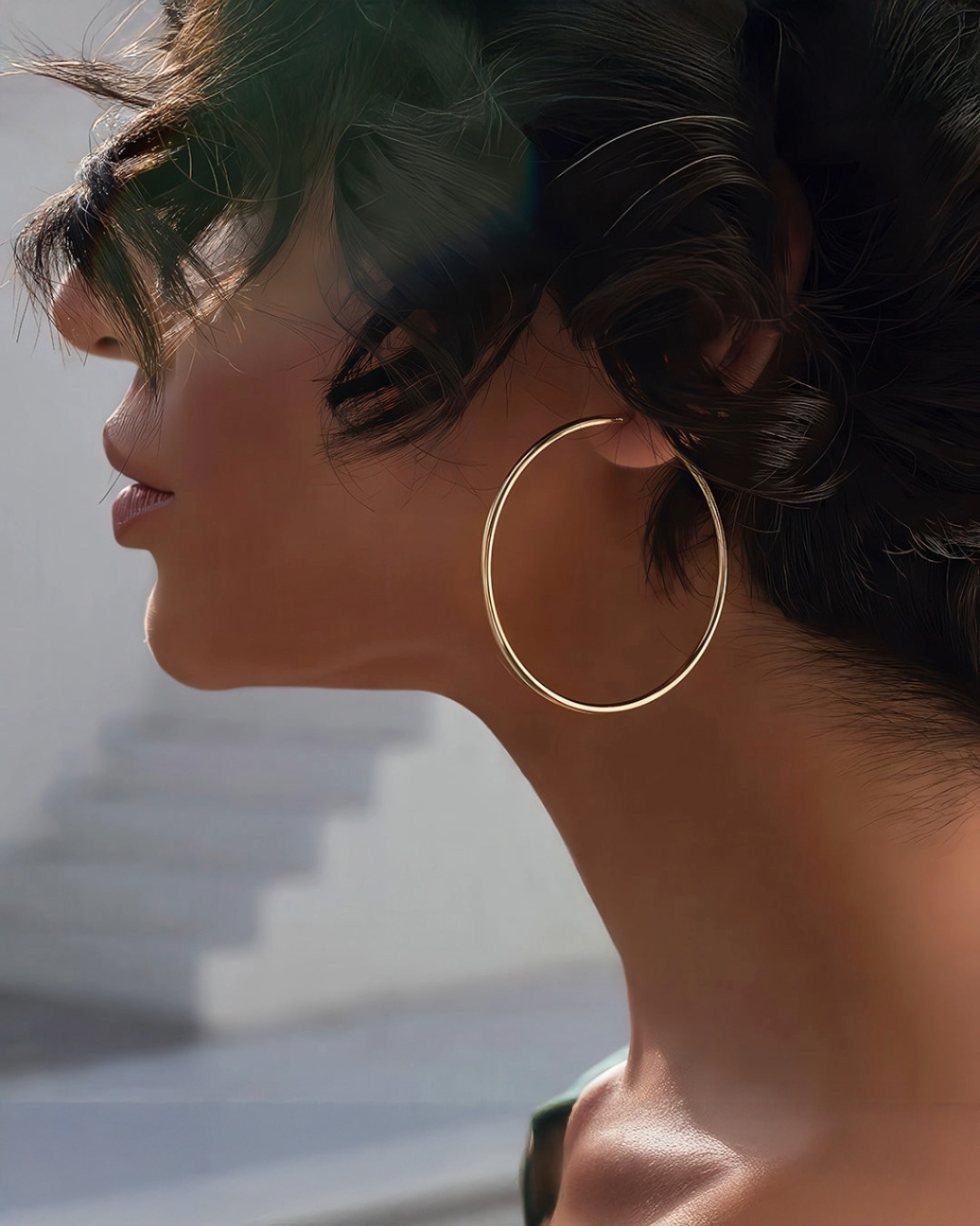 Seitenprofil einer eleganten Frau mit hochgestecktem, dunklem Haar, die einen großen, runden Creolen-Ohrring aus Gold trägt. Weiches Tageslicht betont die Konturen von Gesicht und Hals, der Hintergrund ist ruhig und unscharf.