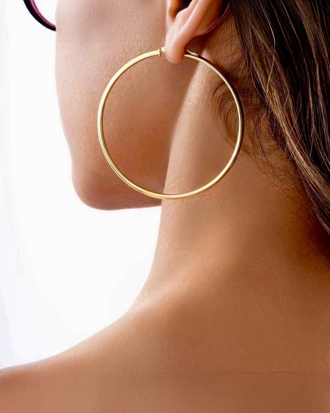 Detailaufnahme eines großen, runden Creolen-Ohrrings aus Gold am Ohr einer Frau. Der Fokus liegt auf der feinen, glänzenden Oberfläche des Ohrrings sowie den eleganten Linien von Kiefer, Hals und Schulter vor hellem, minimalistischem Hintergrund.