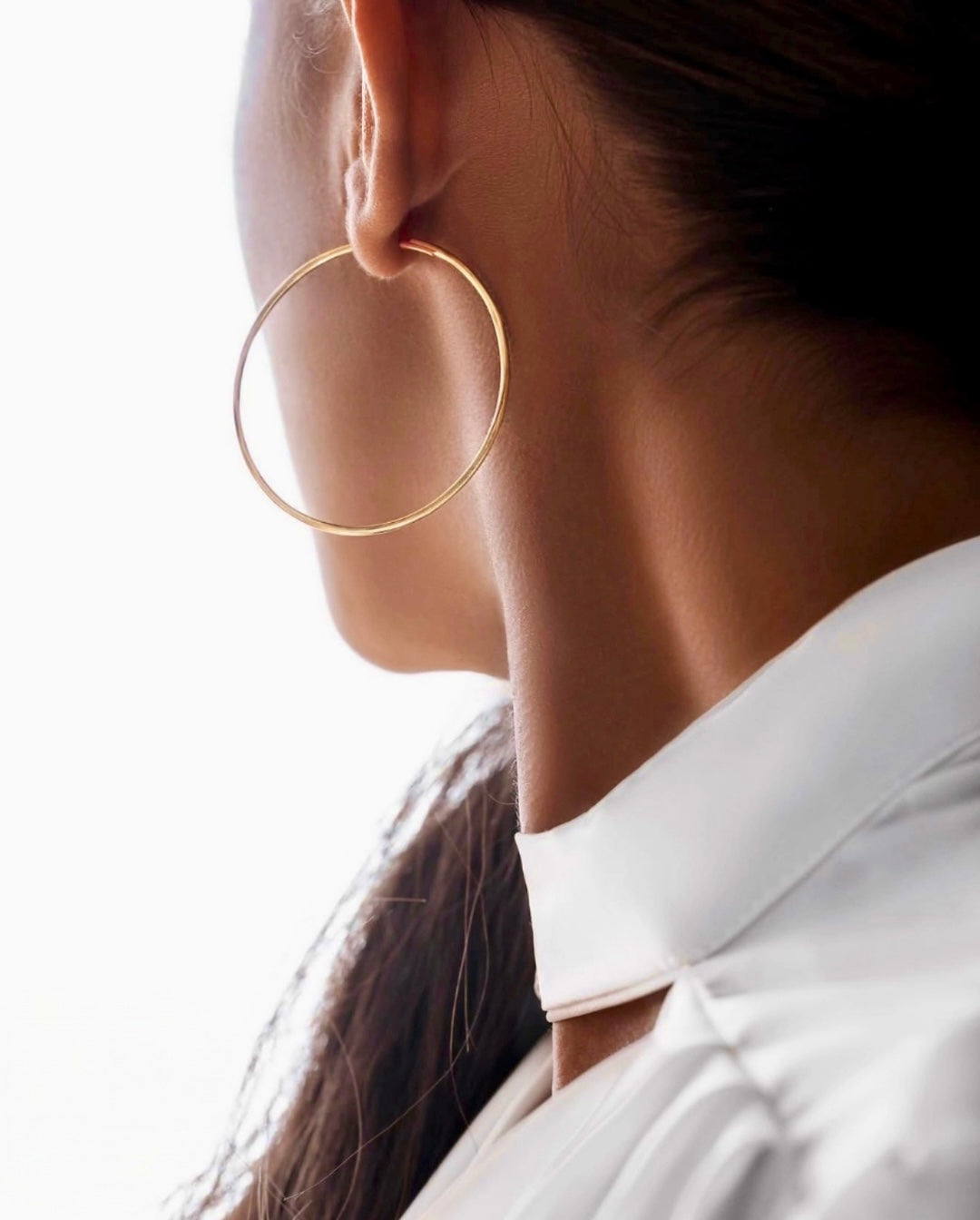 Elegante Nahaufnahme einer Person mit großem, dünnem Creolen-Ohrring in Gold. Das Haar ist zurückgebunden, der Fokus liegt auf Ohr, Hals und Schmuckstück. Die Person trägt ein weißes Oberteil mit hohem Kragen vor hellem, minimalistischem Hintergrund. Die Aufnahme wirkt modern, stilvoll und luxuriös.